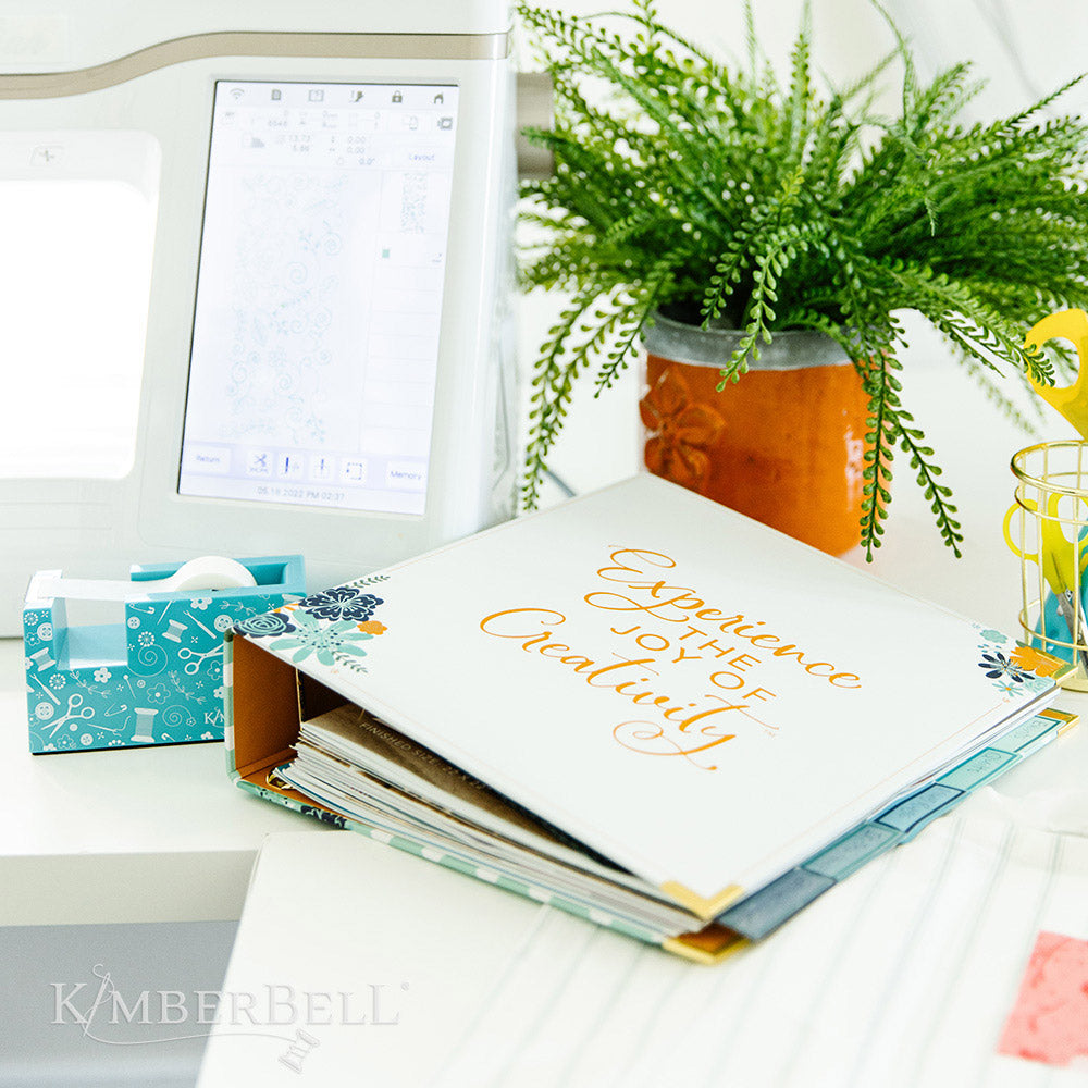 Inspire yourself with the Experience the Joy Binder (KDMR131) by Kimberbell. Each 3" binder includes 5 tab dividers and 2 sheets of stickers, plus gold metal corners to ensure the durability. This photo shows it partially filled while laying on a desk.