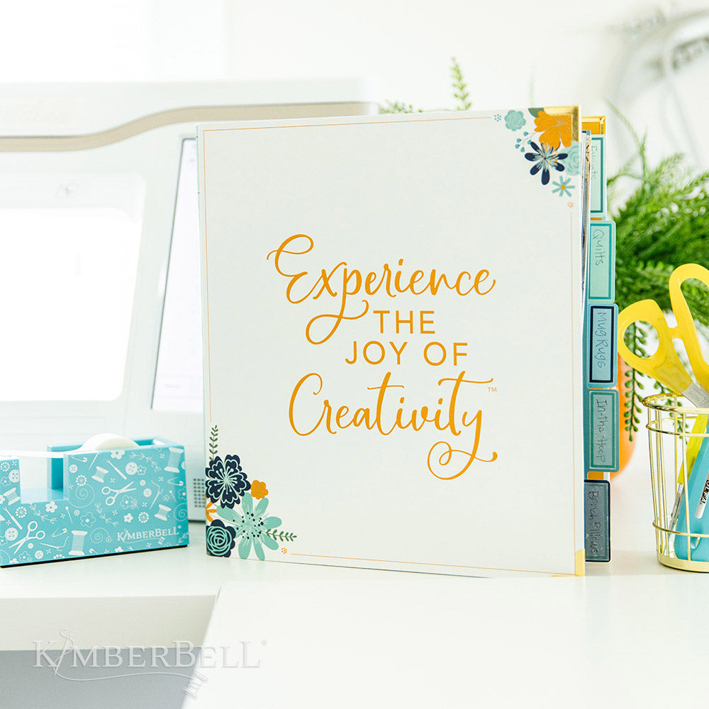 Inspire yourself with the Experience the Joy Binder (KDMR131) by Kimberbell. Each 3" binder includes 5 tab dividers and 2 sheets of stickers, plus gold metal corners to ensure the durability. This binder shows the front standing up on a desk.