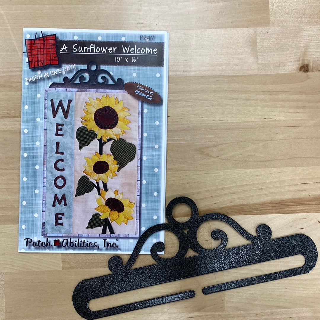 This Welcome banner features lifelike sunflowers right from the garden to brighten any room with A Sunflower Welcome (P247) mini quilt pattern by PatchAbilities. Photo shows the cover of the pattern with the included hanger.