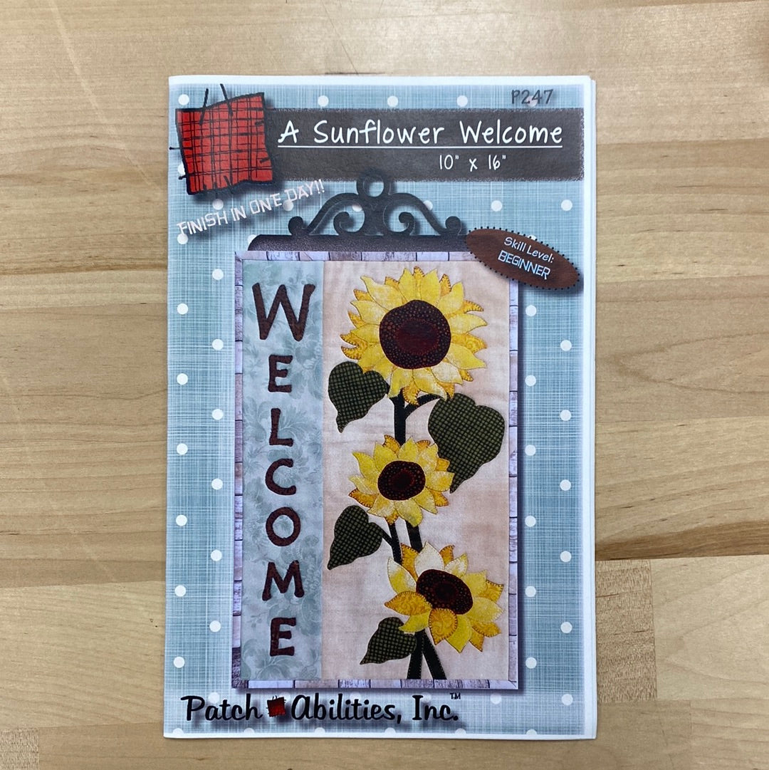 This Welcome banner features lifelike sunflowers right from the garden to brighten any room with A Sunflower Welcome (P247) mini quilt pattern by PatchAbilities. Photo shows the front cover of the pattern.