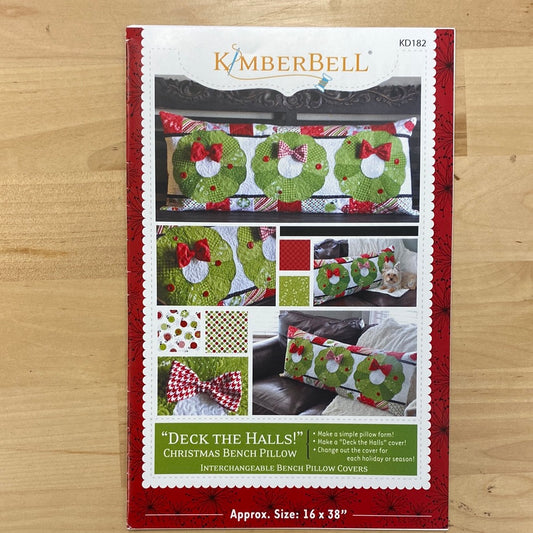 Get festive with our Deck the Halls! Bench Pillow Pattern (KD182) by Kimberbell. Adorned with appliqued wreaths and bows, this pattern includes a pieced border for added charm. Perfect for regular sewing machines, it's the perfect addition to your holiday decor. Photo shows the front cover of the package.