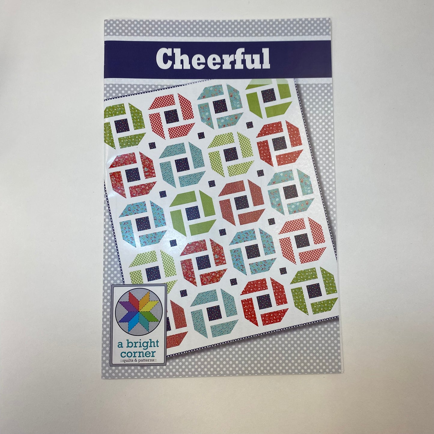 A quilt pattern booklet titled 'Cheerful' by a bright corner, featuring a colorful geometric block pattern on the cover, photographed against a neutral light gray background.