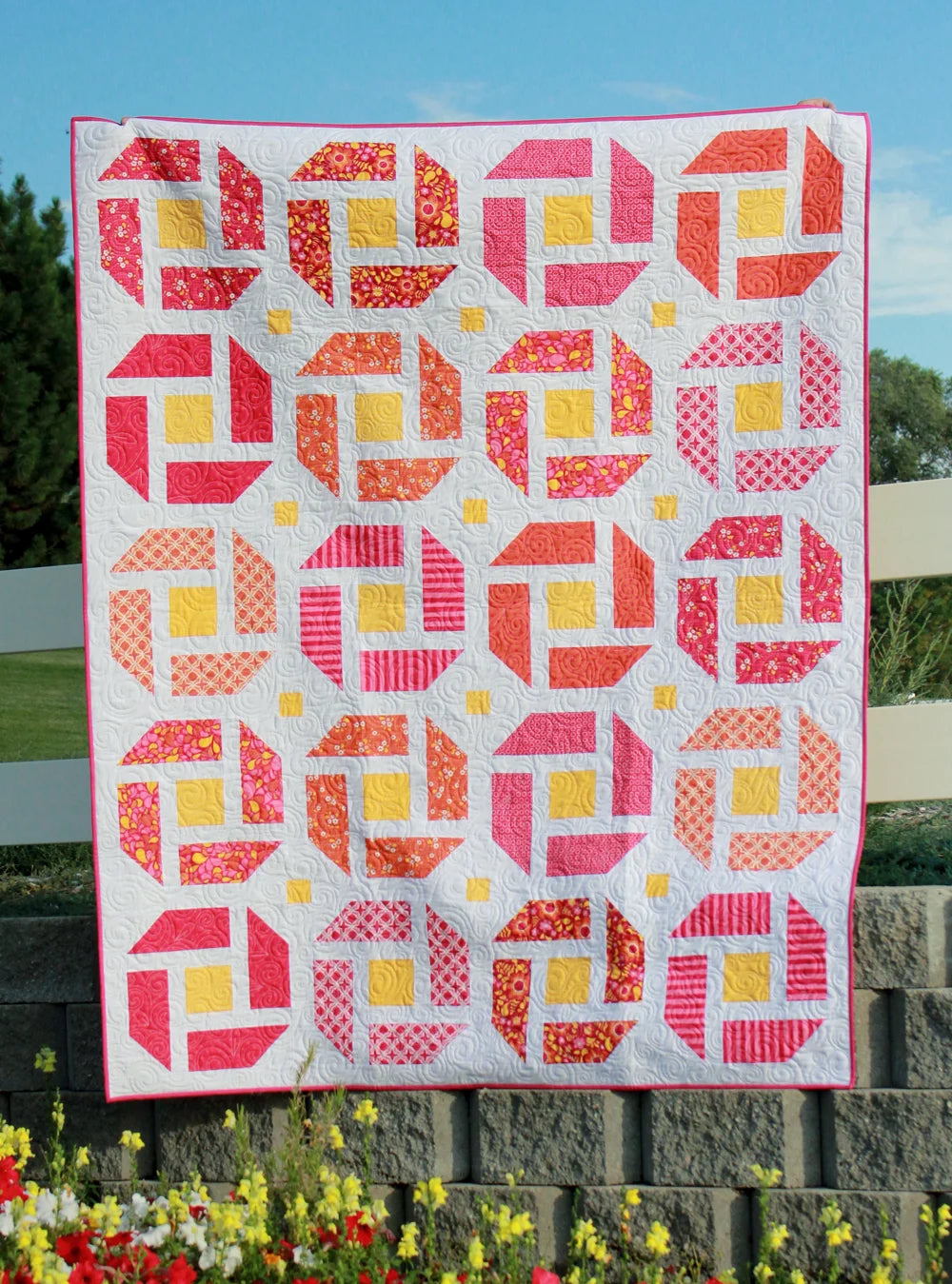 A colorful quilted patchwork blanket or small quilt hanging outdoors against a white fence, featuring repeating geometric blocks in pink, red, orange, and yellow on a white background.