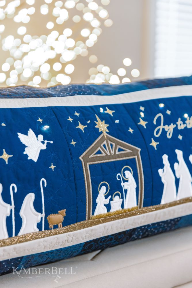 Celebrate the season with Kimberbell’s Nativity Bench Pillow (KD5127)! The holy family is safely ensconced in an applique stable, while Fairy Lights twinkle in the starry night sky. Shepherds and wise men are coming to greet them, a shimmering glitter star and angel guiding their way! Photo shows detail of the Holy Family on the finished pillow.