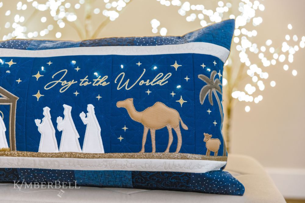 Celebrate the season with Kimberbell’s Nativity Bench Pillow (KD5127)! The holy family is safely ensconced in an applique stable, while Fairy Lights twinkle in the starry night sky. Shepherds and wise men are coming to greet them, a shimmering glitter star and angel guiding their way! Photo shows detail of the wise men and 'Joy to the World' sentiment on the finished pillow.