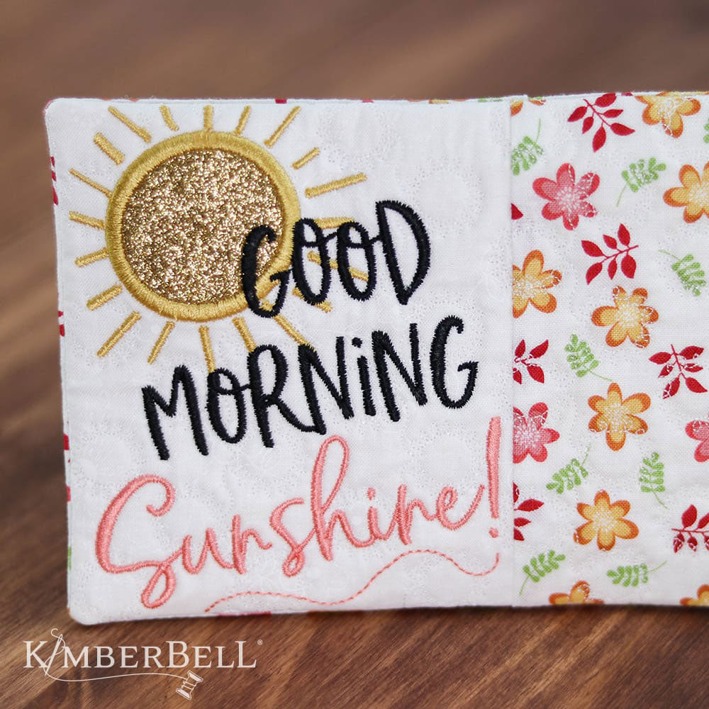 Kimberbell Mug Rug pattern for machine embroidery features small projects with fun sayings to liven up a desk or table. This photo shows the Sunshine pattern stitched out, with a machine applique sun and the words Good Morning Sunshine.