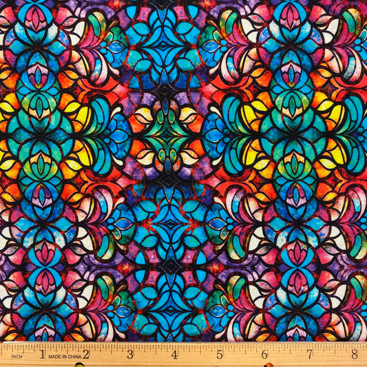 A close-up of a colorful patterned fabric with a repeating stained-glass style floral and geometric design in blue, teal, yellow, orange, pink, purple, and black. A wooden ruler is visible along the bottom edge for scale.