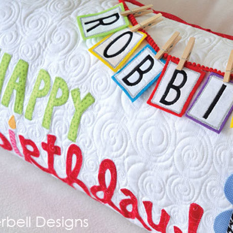 The Happy Birthday Bench pillow patten for machine embroidery by Kimberbell features all the embroidery files for making. The pillow features a topsy, turvy cake, Happy Birthday, and interchangeable name banner for celebrating the special someone. The photo shows the detail of the printable letters that can be attached to the pillow with mini clothes pins.