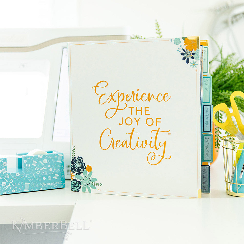 Inspire yourself with the Experience the Joy Binder (KDMR131) by Kimberbell.  Each 3" binder includes 5 tab dividers and 2 sheets of stickers, plus gold metal corners to ensure the durability. This binder shows the front standing up on a desk.