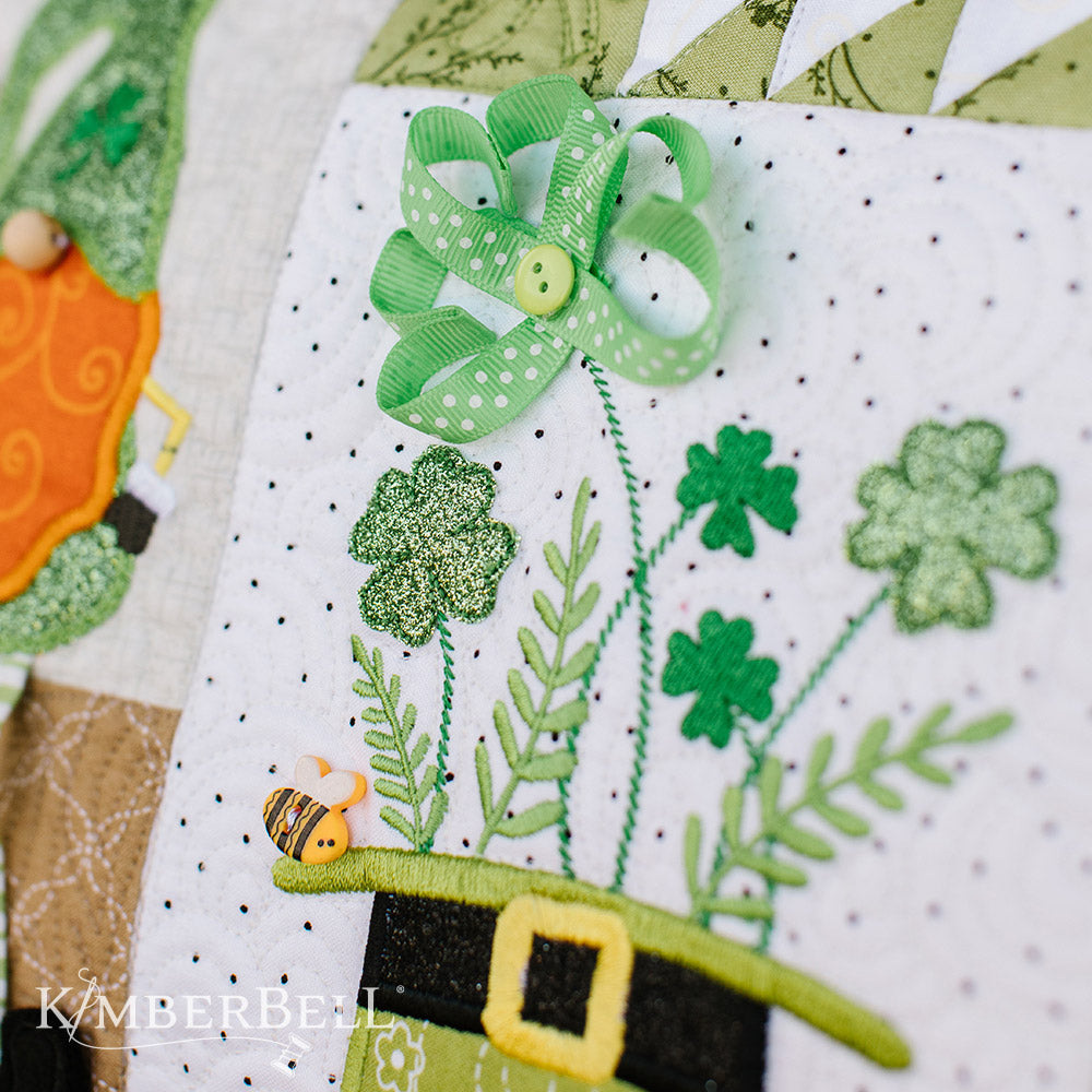 With wee folk, wishes, and decorative stitches, the Kimberbell ​Luck o’ the Gnome: St. Patrick’s Day Bench Pillow for machine embroidery (KD587)​ has “all the joy your heart can hold! Photo shows the hat detail, including a ribbon shamrock and applique glitter shamrocks.