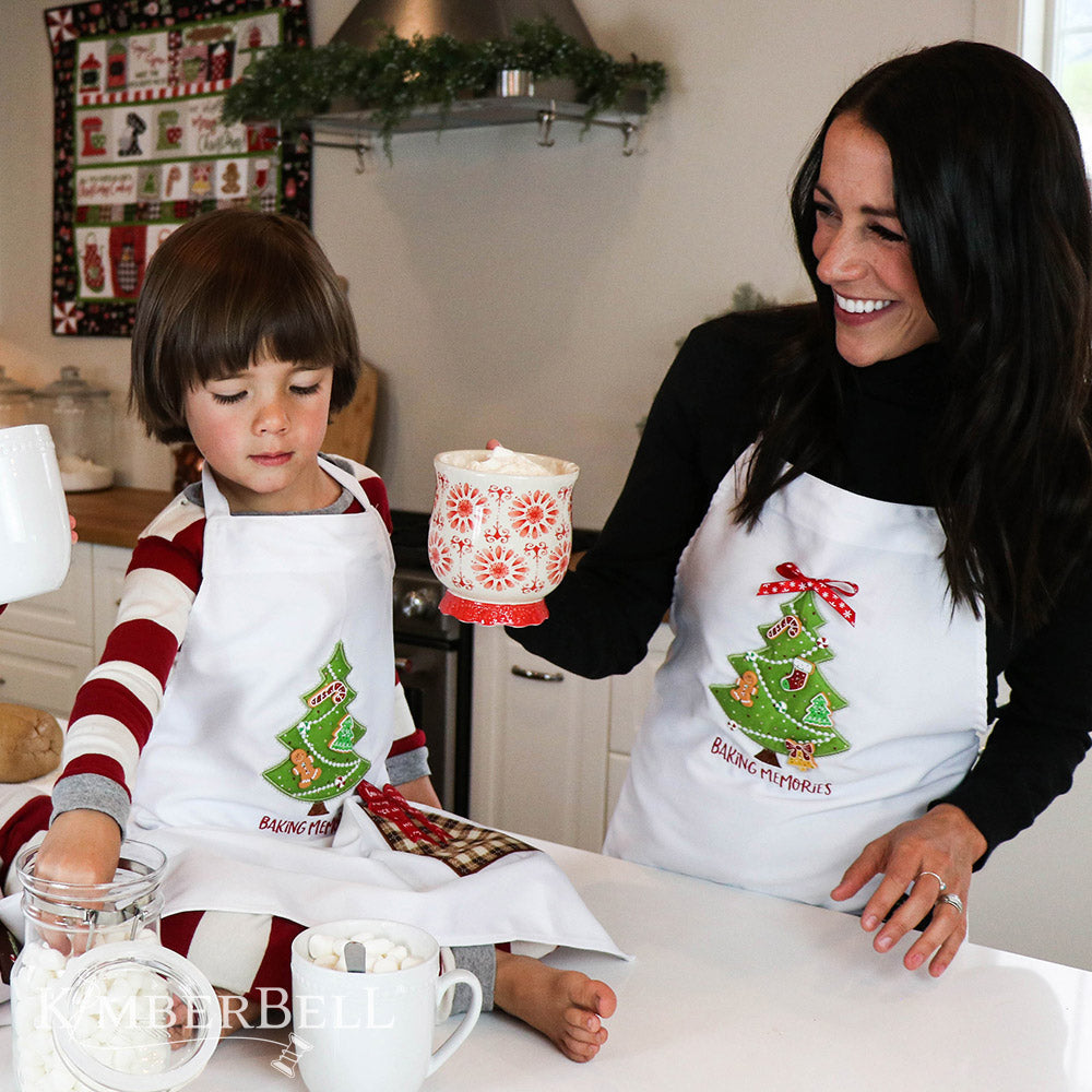 We “Whisk” You a Merry Christmas (KD806) is Kimberbell’s feature quilt and holiday tribute to sweet Christmas confections and even sweeter traditions and memories. Dimensional elements include flexible foam whipped cream, marshmallow poms, a clear vinyl cake dome, applique glitter countertop mixers, and more! Picture shows child and mother in embroidered aprons with Christmas tree.