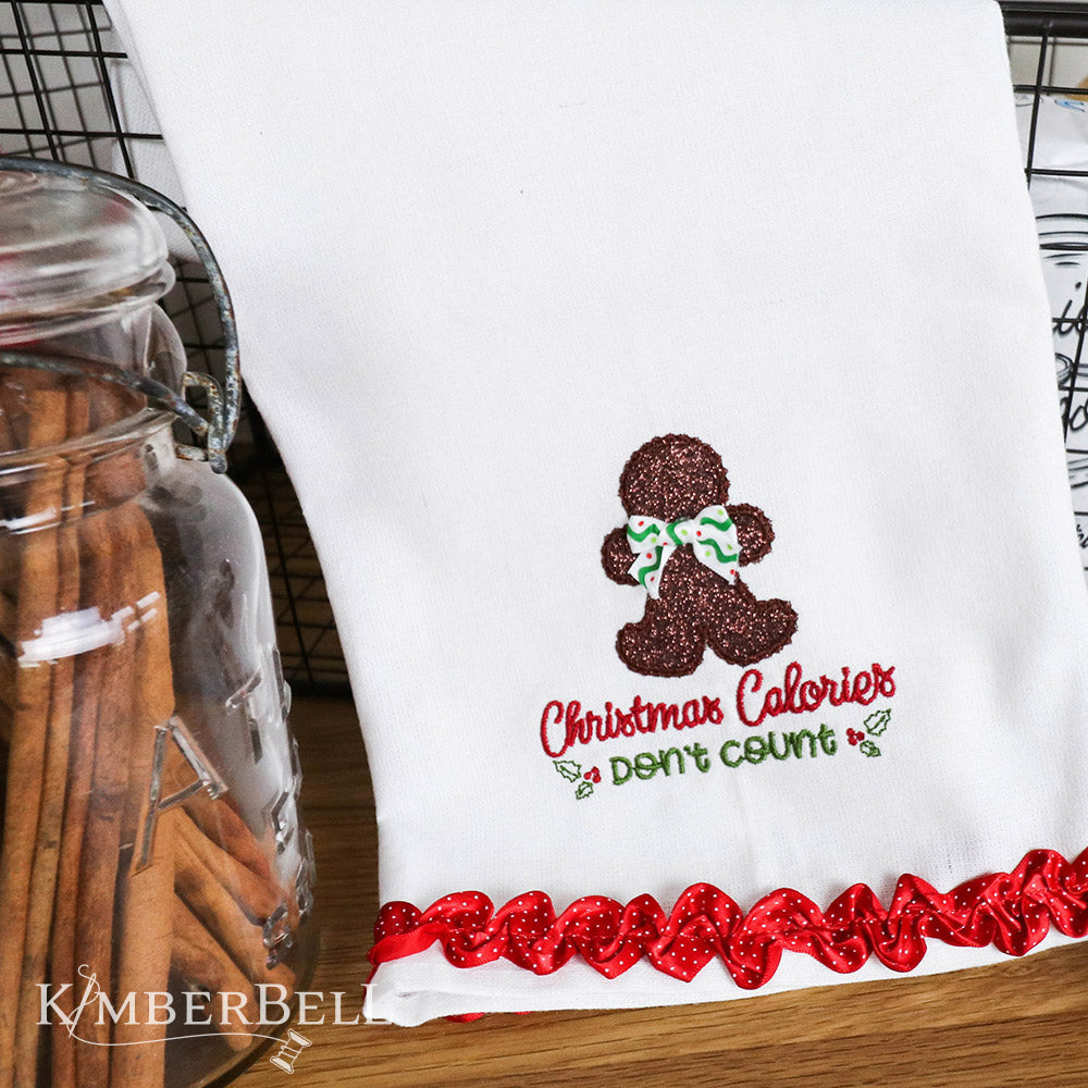 We “Whisk” You a Merry Christmas (KD806) is Kimberbell’s feature quilt and holiday tribute to sweet Christmas confections and even sweeter traditions and memories. Dimensional elements include flexible foam whipped cream, marshmallow poms, a clear vinyl cake dome, applique glitter countertop mixers, and more! Picture shows tea towel embroidered with gingerman and ruffle embellishment.